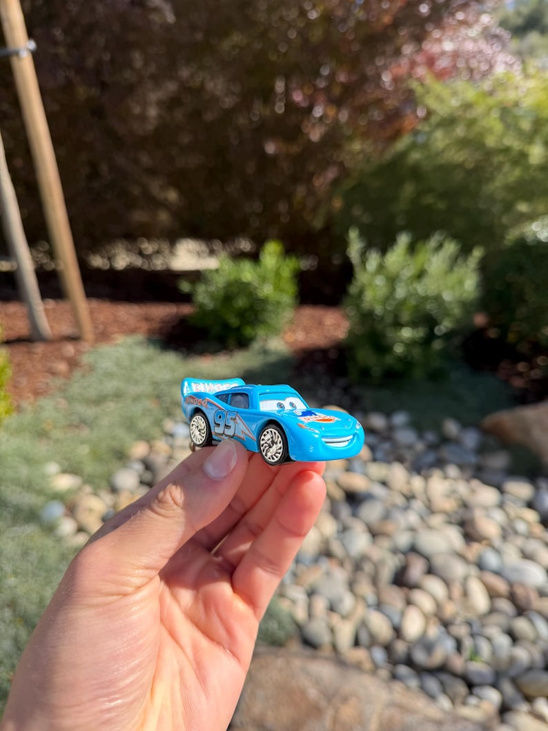Może przedstawiać: Jasnoniebieski zabawkowy samoch&oacute;d wyścigowy z numerem "95" z boku. Samoch&oacute;d ma uśmiechniętą twarz i spojler. Koła są srebrne. Samoch&oacute;d jest trzymany w dłoni na tle zieleni i skał.