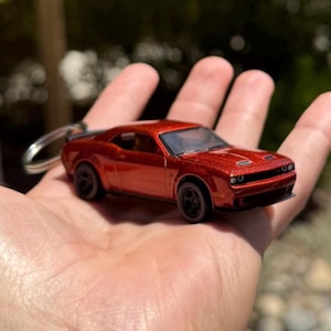 以下が含まれることがあります： ミニチュアの赤いスポーツカーのキーホルダー。おもちゃの車は黒い車輪と、スポイラーやボンネットの通気孔など、詳細なデザインが特徴です。車は手で持たれており、そのサイズとメタリックな仕上がりを示しています。
