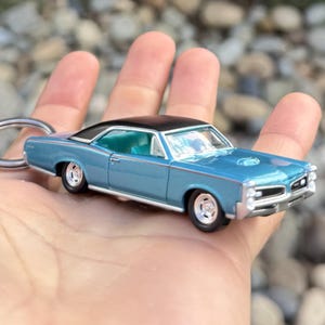 May include: A miniature blue classic car with a black roof, chrome accents, and silver wheels. The model car is held in a hand, showcasing its details. The car has a keychain ring attached.