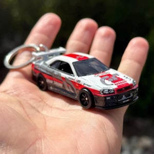 Könnte beinhalten: Ein Miniatur-Schlüsselanhänger mit einem silbernen, roten und weißen Nissan Skyline GT-R R34 Modellauto. Das Auto hat schwarze Räder, einen Heckspoiler und die Aufschrift "GRAN TURISMO" und "NISSAN".