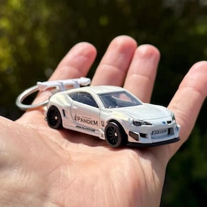 Peut inclure: Une petite voiture blanche avec des roues noires et un aileron arrière, tenue dans une main. La voiture porte le mot "PANDEM" sur le côté et est attachée à un porte-clés. C'est une réplique miniature d'une voiture de sport.