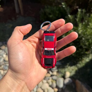 May include: Red die-cast model car keychain with white and black accents. The keychain features the Castrol logo and Firestone branding. The car is a miniature replica of a classic race car, attached to a silver keyring.