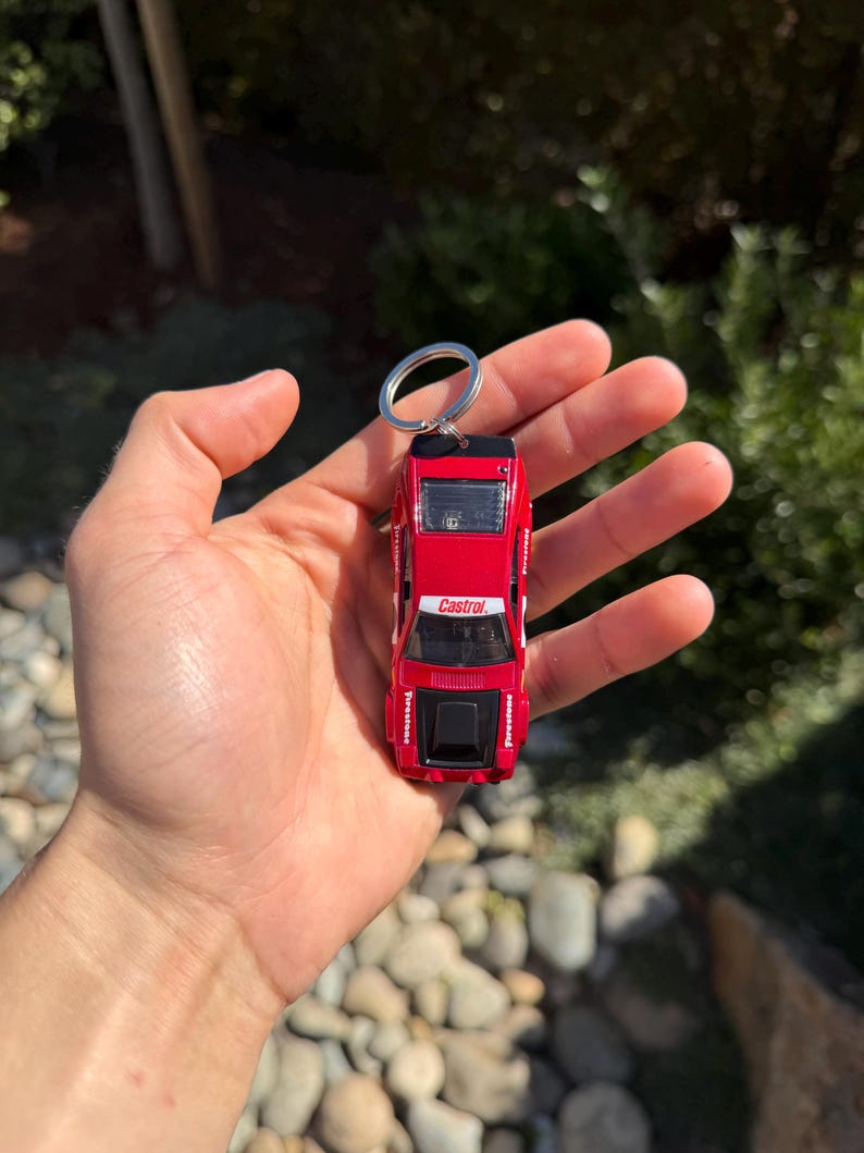 May include: Red die-cast model car keychain with white and black accents. The keychain features the Castrol logo and Firestone branding. The car is a miniature replica of a classic race car, attached to a silver keyring.