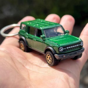 May include: A green die-cast model of a Bronco SUV with a keychain attachment. The miniature vehicle features black tires with gold rims and the word "BRONCO" on the front. The model is metallic green.