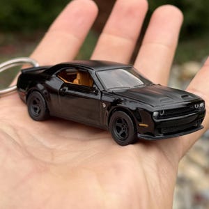 以下が含まれることがあります： 詳細なデザインのミニチュア黒スポーツカーキーホルダー。車は黒いホイール、フードスクープ、タンの内装を備えています。キーホルダーは手で持たれています。
