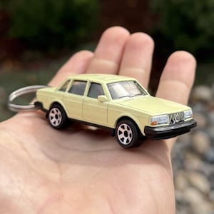 May include: A miniature yellow sedan keychain with black wheels and a silver key ring. The toy car has a black front grill and headlights. The car is approximately 7.6 cm long.
