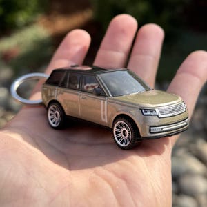 May include: A miniature gold SUV keyring with a black roof and silver wheels. The toy car is held in an open hand, showcasing its detailed design. The keyring ring is silver.