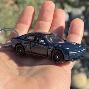 以下が含まれることがあります： 濃い青色のダイキャスト製モデルカーキーホルダー。 ミニカーは黒いホイールと黒い屋根が付いています。 キーホルダーは手で持っており、背景はぼやけています。