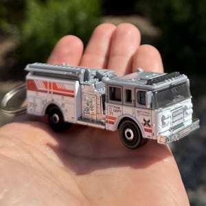 May include: A miniature white and gray fire truck toy with red accents, held in a hand. The toy truck has the words "FIRE DEPT 71" and "MATCHBOX COUNTY" printed on it. The toy is attached to a silver keychain.
