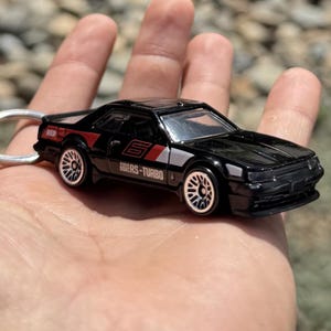 Può includere: Un'auto giocattolo nera con dettagli rossi e argento, con le scritte "RED" e "RS-TURBO". Il veicolo in miniatura ha ruote bianche e un portachiavi. L'auto è tenuta in una mano.