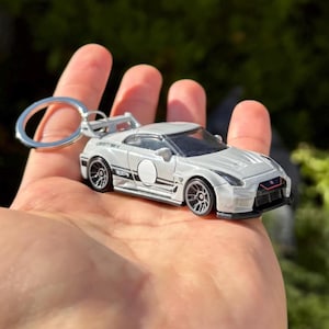 May include: A silver die-cast car keychain with a spoiler and black wheels. The car has a white stripe and the text "LIBERTY WALK". The keychain ring is silver.