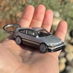 Peut inclure: Porte-clés avec une voiture miniature argentée. La voiture miniature a un design à hayon avec des garnitures noires et des roues grises. L'anneau du porte-clés est argenté.