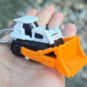 May include: A small white and orange bulldozer keyring. The toy bulldozer has black tracks, an orange blade, and the text "RANEC EQUIPMENT". The keyring is made of metal and plastic.
