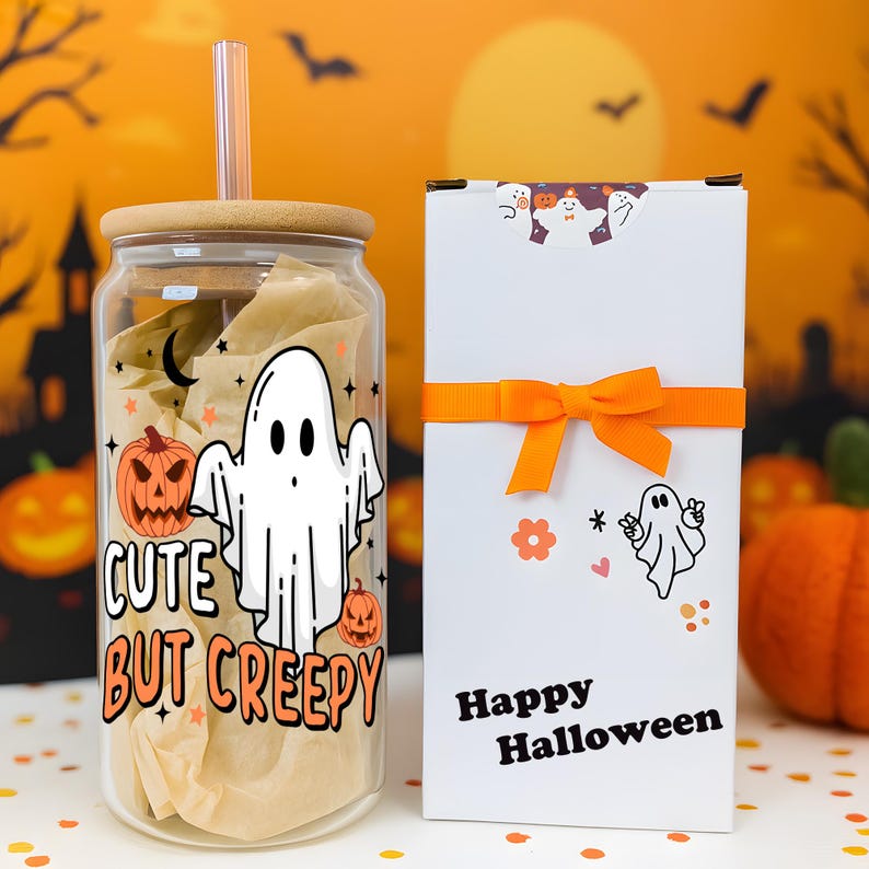 May include: A clear glass tumbler with a bamboo lid and straw, featuring a white ghost, orange pumpkins, and the text "CUTE BUT CREEPY". Beside it is a white rectangular gift box with an orange ribbon, a ghost sticker, and "Happy Halloween" text.