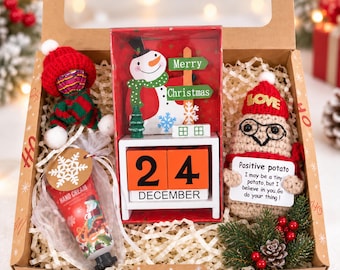 May include: A gift box containing a red and green crocheted doll in a hat, a tube of hand cream with a snowflake tag, a wooden perpetual calendar with orange blocks showing 24 December and a snowman sign, and a crocheted potato doll with a sign.