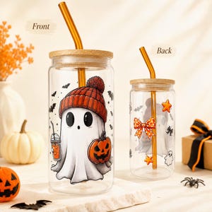 May include: Two clear glass tumblers with bamboo lids and amber glass straws. The larger tumbler features a ghost wearing an orange beanie holding a jack-o'-lantern. The smaller tumbler shows a ghost with an orange polka dot bow and stars.