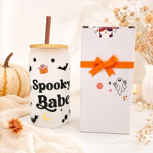 May include: A frosted glass tumbler with a bamboo lid and brown straw, featuring black text reading Spooky Babe, black bats, polka dots, a yellow crescent moon, and a pumpkin wearing a pink cowboy hat, next to a white gift box with an orange ribbon.