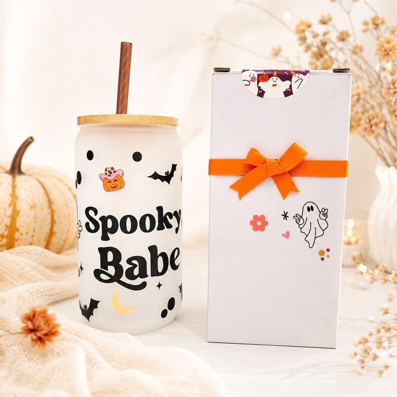 May include: A frosted glass tumbler with a bamboo lid and brown straw, featuring black text reading Spooky Babe, black bats, polka dots, a yellow crescent moon, and a pumpkin wearing a pink cowboy hat, next to a white gift box with an orange ribbon.