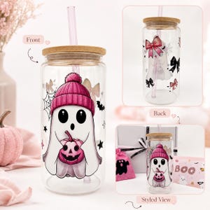May include: A clear glass tumbler with a bamboo lid and a pink straw. The front features a ghost wearing a pink beanie and holding a pink pumpkin. The back displays pink bows and spiderwebs. The design includes black stars and bats.