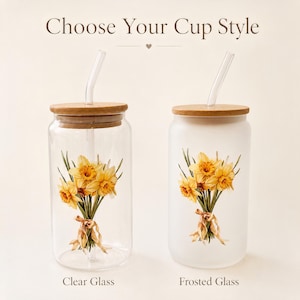 May include: Two glass cups with bamboo lids and glass straws, featuring a bouquet of yellow daffodils tied with a ribbon. The left cup is clear glass, and the right cup is frosted glass. The text above reads Choose Your Cup Style.