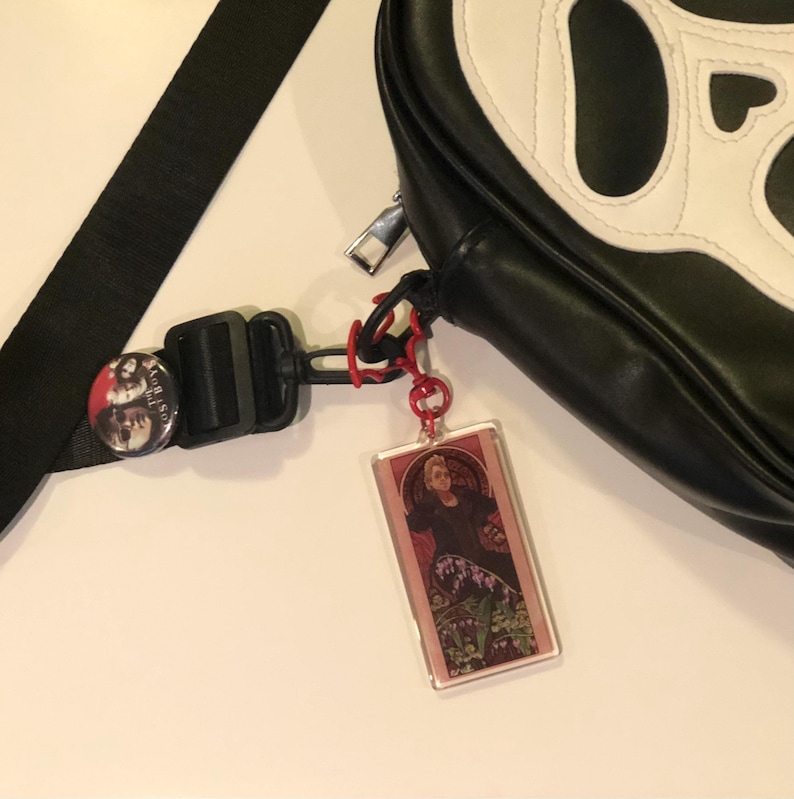 May include: A black and white bag with a skull design. The bag has a red and black strap with a button and a clear plastic charm with a picture of a person.