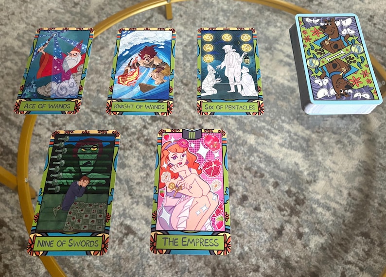 K&ouml;nnte beinhalten: Ein Tarotkartenspiel mit farbenfrohen Illustrationen. Die Karten zeigen Bilder von Stab-Ass, Ritter der St&auml;be, Sechs der Pentakel, Neun der Schwerter und Die Kaiserin. Ein Kartenspiel mit Scooby Doo-Design ist ebenfalls vorhanden.