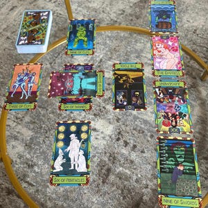 K&ouml;nnte beinhalten: Ein Tarotkartenspiel mit farbenfrohen Illustrationen, darunter Karten mit der Aufschrift "Nine of Cups", "Three of Cups" und "Six of Wands". Die Karten sind auf einem Glastisch mit goldenem Metallrahmen angeordnet.