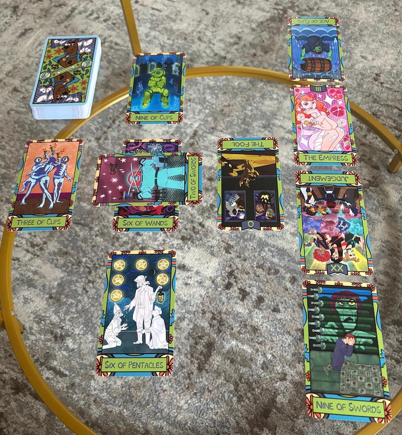 K&ouml;nnte beinhalten: Ein Tarotkartenspiel mit farbenfrohen Illustrationen, darunter Karten mit der Aufschrift "Nine of Cups", "Three of Cups" und "Six of Wands". Die Karten sind auf einem Glastisch mit goldenem Metallrahmen angeordnet.