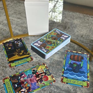 K&ouml;nnte beinhalten: Ein Tarotkartenspiel mit farbenfrohen Illustrationen, darunter "The Fool", "Judgement" und "Ace of Cups". Die Karten werden mit einer wei&szlig;en Box auf einem Glastisch pr&auml;sentiert. Die Kartenr&uuml;ckseiten weisen ein sich wiederholendes Muster auf.