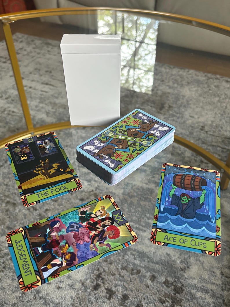 K&ouml;nnte beinhalten: Ein Tarotkartenspiel mit farbenfrohen Illustrationen, darunter "The Fool", "Judgement" und "Ace of Cups". Die Karten werden mit einer wei&szlig;en Box auf einem Glastisch pr&auml;sentiert. Die Kartenr&uuml;ckseiten weisen ein sich wiederholendes Muster auf.