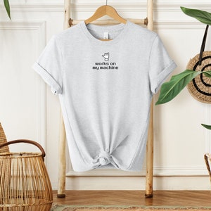 May include: A light grey t-shirt with a black graphic of a hand giving a thumbs up and the text "works on my machine".