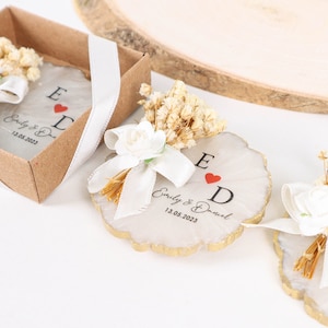 May include: Two white wooden round ornaments with gold trim, decorated with dried flowers and a white ribbon. The ornaments have the letters "E" and "D" with a red heart in between, and the text "Emily & Daniel" and a date.