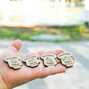 May include: Four wooden baby onesie-shaped ornaments with the text "Welcome Baby" and a baby-related icon. Each ornament has a name and date, such as "Michael 22.07.2024", "James 22.07.2004", and "Sophia 22.07.2024".