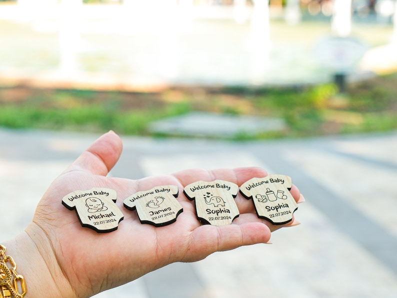 May include: Four wooden baby onesie-shaped ornaments with the text "Welcome Baby" and a baby-related icon. Each ornament has a name and date, such as "Michael 22.07.2024", "James 22.07.2004", and "Sophia 22.07.2024".