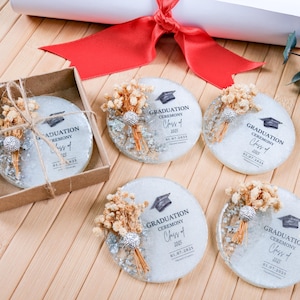 May include: Graduation-themed coasters with dried floral accents. Each coaster features the text "GRADUATION CEREMONY Class of 2025" and the date 01.07.2025. One coaster is packaged in a small brown box, tied with twine.