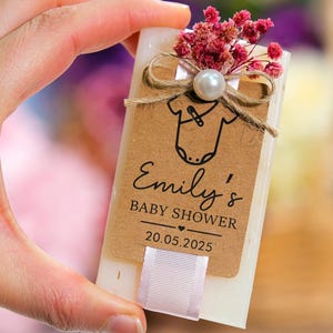 May include: A baby shower favor with a kraft paper tag that reads "Emily's BABY SHOWER 20.05.2025". The tag features a onesie graphic and is adorned with a pearl, twine, and dried pink flowers. A white ribbon is attached.