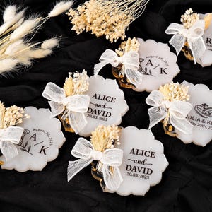 May include: White, scalloped-edge coasters with personalized text and dates, decorated with dried flowers and white bows. The coasters feature names and dates, perfect for wedding favors. The background is black.