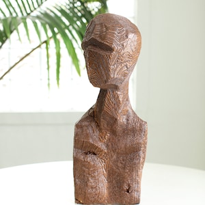 Puede incluir: Una escultura de madera de la cabeza y los hombros de una persona. La escultura está tallada con una superficie rugosa y texturizada y está pintada de un color marrón oscuro.