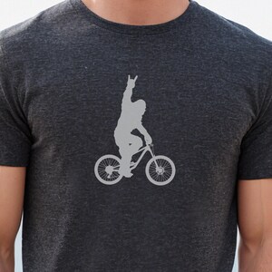 May include: Dark gray t-shirt featuring a gray silhouette of a person riding a bicycle and raising their hand in a rock-and-roll gesture. The bicycle has large wheels and is designed for mountain biking. The shirt is made of a soft, comfortable material.