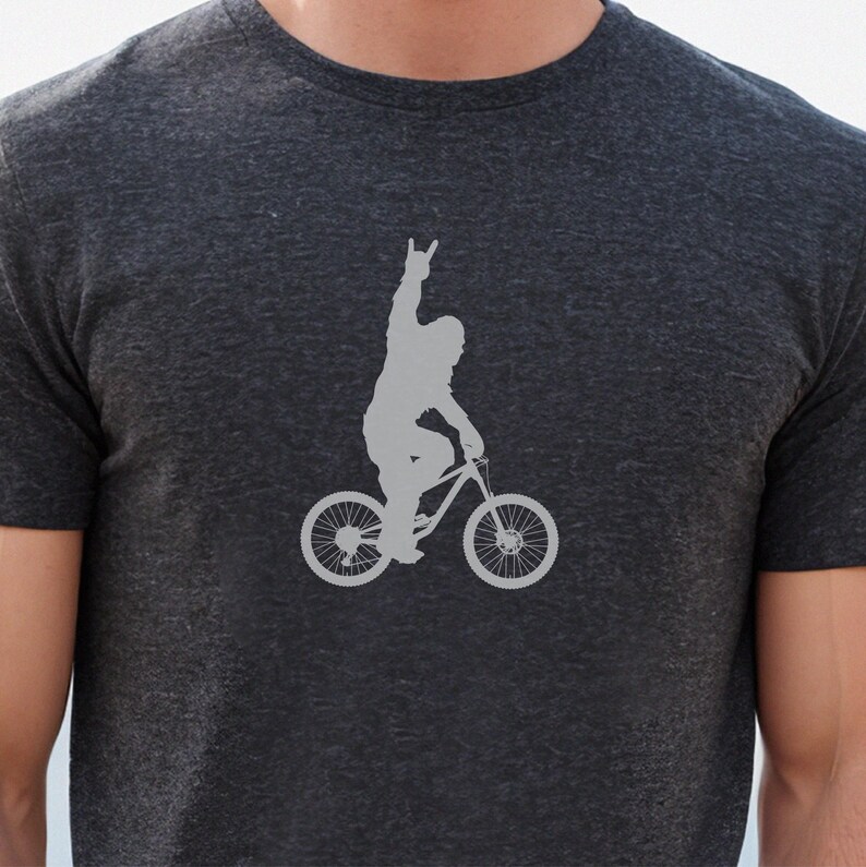 May include: Dark gray t-shirt featuring a gray silhouette of a person riding a bicycle and raising their hand in a rock-and-roll gesture. The bicycle has large wheels and is designed for mountain biking. The shirt is made of a soft, comfortable material.