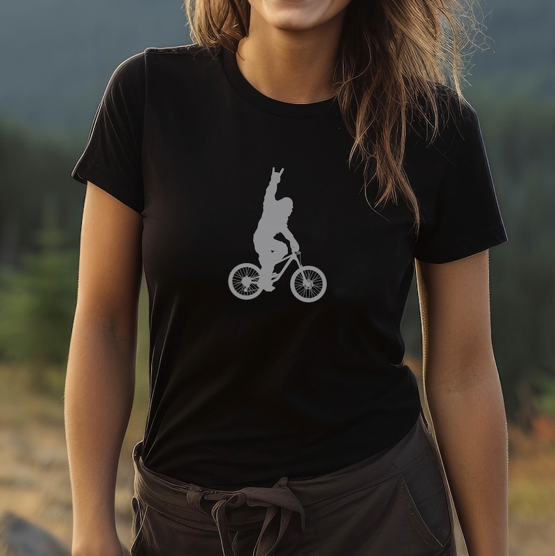 May include: Black t-shirt featuring a gray silhouette of a person riding a bicycle with one arm raised in a rock-on gesture. The bicycle has large tires. The shirt is made of a soft material.