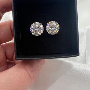 May include: Two round, clear gemstone stud earrings displayed in a black box. The earrings are set in a silver metal and reflect light, creating a sparkling effect. The box is held against a white, silky background.