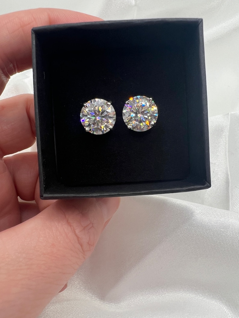 May include: Two round, clear gemstone stud earrings displayed in a black box. The earrings are set in a silver metal and reflect light, creating a sparkling effect. The box is held against a white, silky background.