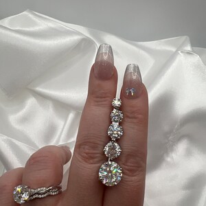 May include: A hand displays a silver ring and a dangling earring with multiple round, clear gemstones. The ring features a large, round gemstone with a twisted band. The earring has a series of graduated stones, culminating in a large, round stone.