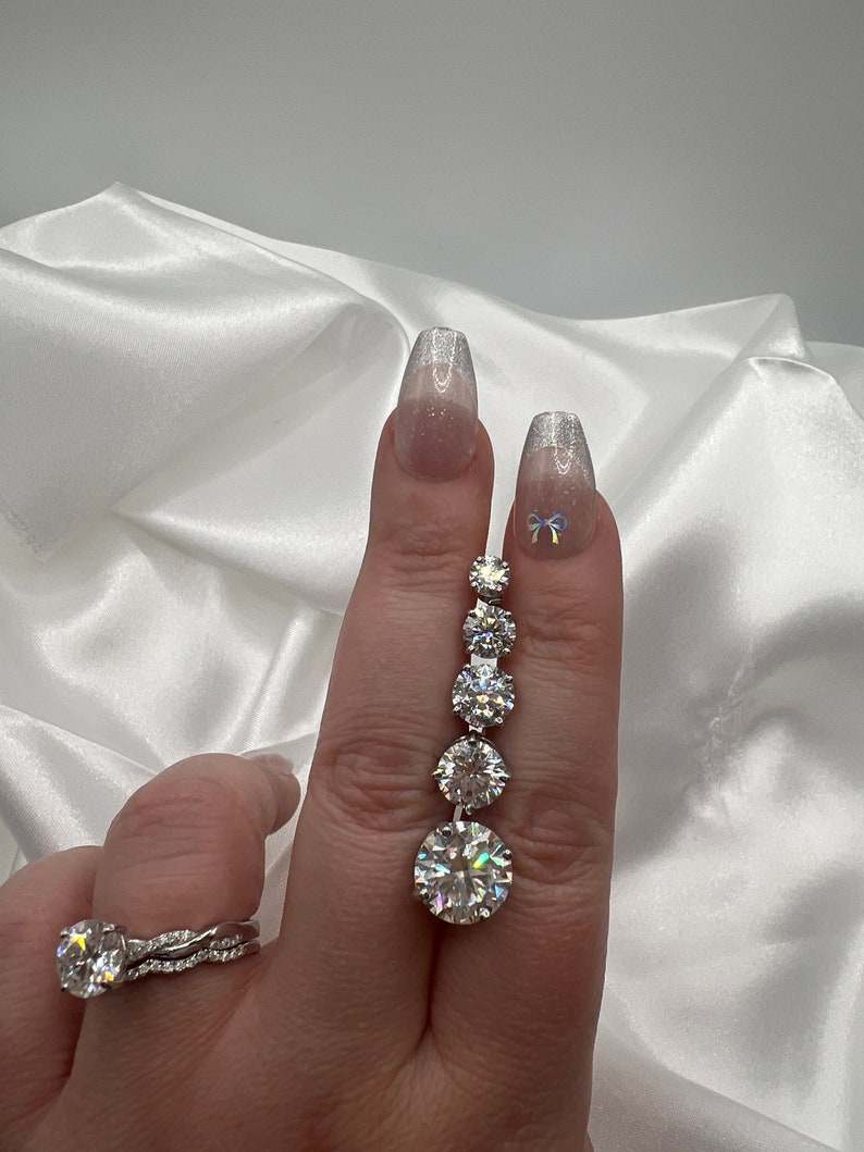 May include: A hand displays a silver ring and a dangling earring with multiple round, clear gemstones. The ring features a large, round gemstone with a twisted band. The earring has a series of graduated stones, culminating in a large, round stone.