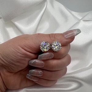 May include: Two round, clear gemstone stud earrings. The earrings are held against a white satin background. The gemstones sparkle and reflect light. The earrings are a classic jewelry item.
