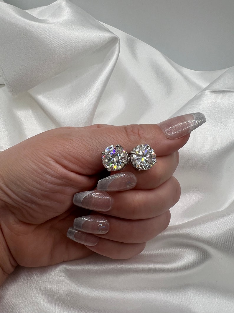 May include: Two round, clear gemstone stud earrings. The earrings are held against a white satin background. The gemstones sparkle and reflect light. The earrings are a classic jewelry item.
