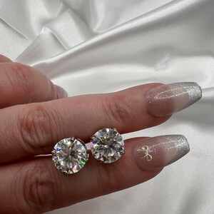 May include: Two round, clear gemstone stud earrings. The earrings are held between fingers against a white satin background. The gemstones sparkle, reflecting light. The nails are painted with a silver glitter design.