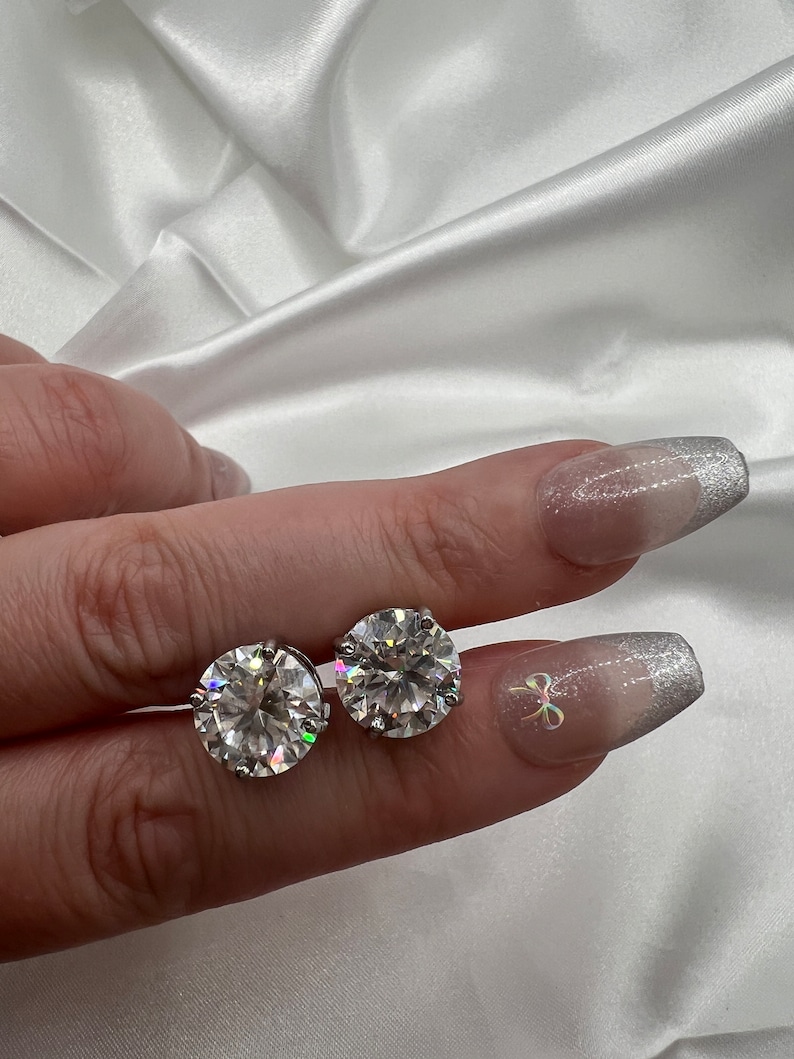 May include: Two round, clear gemstone stud earrings. The earrings are held between fingers against a white satin background. The gemstones sparkle, reflecting light. The nails are painted with a silver glitter design.