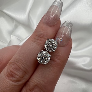 May include: A pair of round, brilliant-cut diamond stud earrings. The diamonds are set in a silver-colored metal and sparkle brightly. The earrings are displayed against a white, silky background.